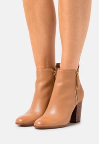 Stivaletti alla caviglia in pelle color tan con finitura lucida, punte arrotondate e dettaglio con zip laterale. I tacchi a blocco sono in legno testurizzato marrone scuro.