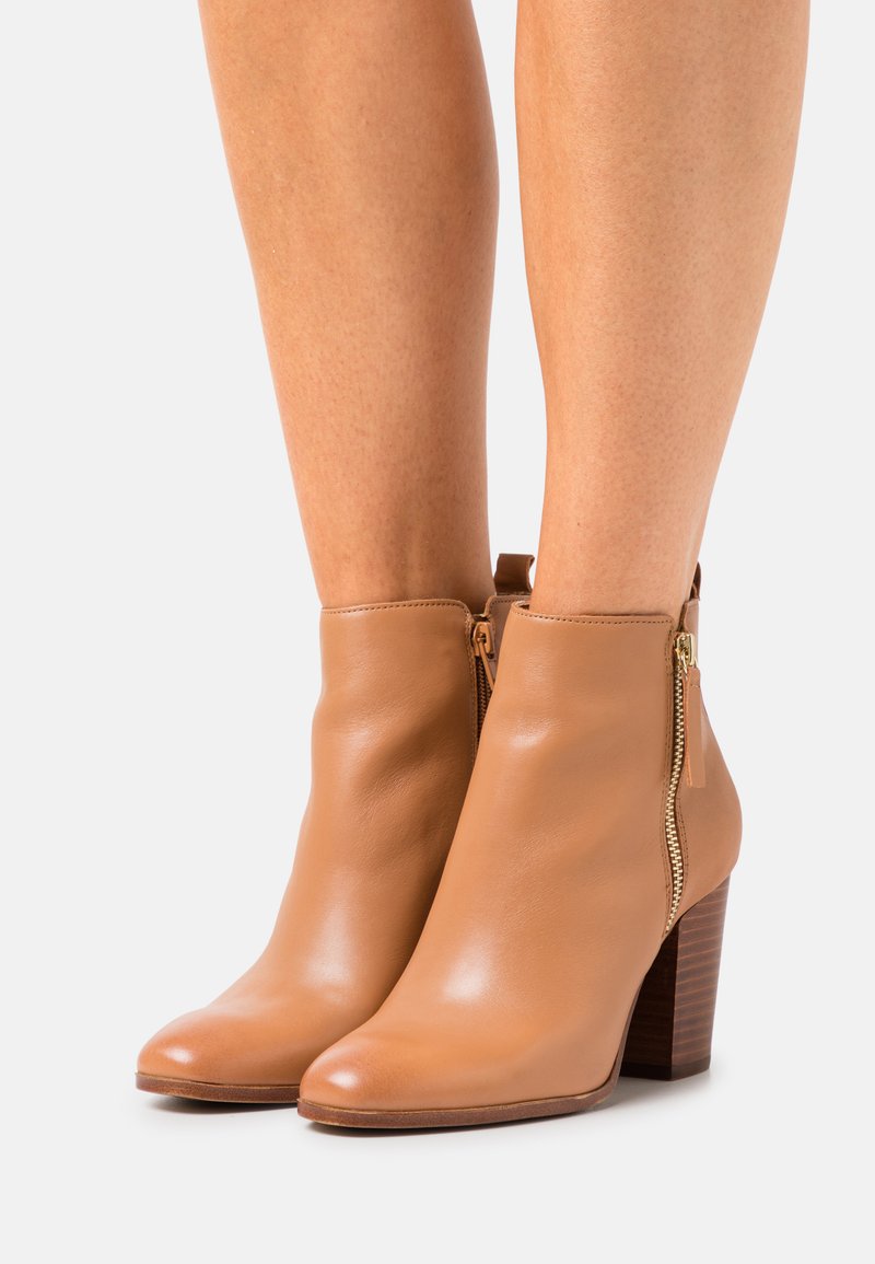 Stivaletti alla caviglia in pelle color tan con finitura lucida, punte arrotondate e dettaglio con zip laterale. I tacchi a blocco sono in legno testurizzato marrone scuro.