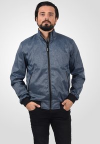 Blå denimjakke i stil med stående krage, glidelås foran og sidelommer. Har svarte mansjetter og en teksturert overflate.