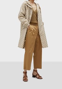 Kvinde iført beige ternet hættetrøje, brun V-hals top, vidbenede brune bukser og brune sandaler med hæl og remme.