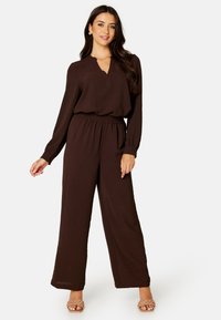 Bruine jumpsuit met lange mouwen, elastische tailleband, V-hals en wijde benen. Glad textuur, gecombineerd met lichtgekleurde sandalen.