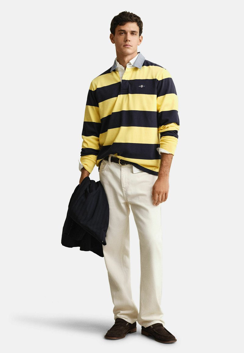 Mann steht und trägt ein gelb-navy gestreiftes Rugbyhemd, weiße Hose, braune Schuhe, hält eine dunkle Jacke in seiner linken Hand.