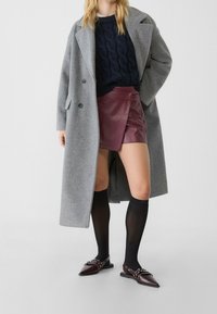 Cappotto di lana grigio, maglione navy a trecce, gonna avvolgente in pelle bordeaux, calze nere a ginocchio e scarpe con punta a punta con dettagli in argento.