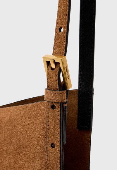 Bandoulière de sac en daim marron avec boucle rectangulaire dorée gravée "AllSaints" et une lanière en cuir noir visible derrière.