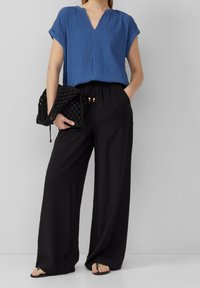 Top blu texture con scollatura a V, abbinato a pantaloni neri larghi con cintura elastica e dettagli in perline di legno, mentre tiene una borsa nera.