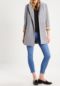 Harmaa villakangas takki, jossa on muotoiltu design, beige hihansuuviimeistely, kaksi vetoketjullista taskua ja hieno kiilto. Yhdistetty sinisiin kapeisiin farkkuihin ja mustiin loafereihin.