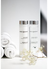 Twee cilindrische flessen met het label 'Lait Végétal' en 'Lotion Végétale' met zilveren doppen; omgeven door netjes opgerolde witte handdoeken en delicate bloemen.