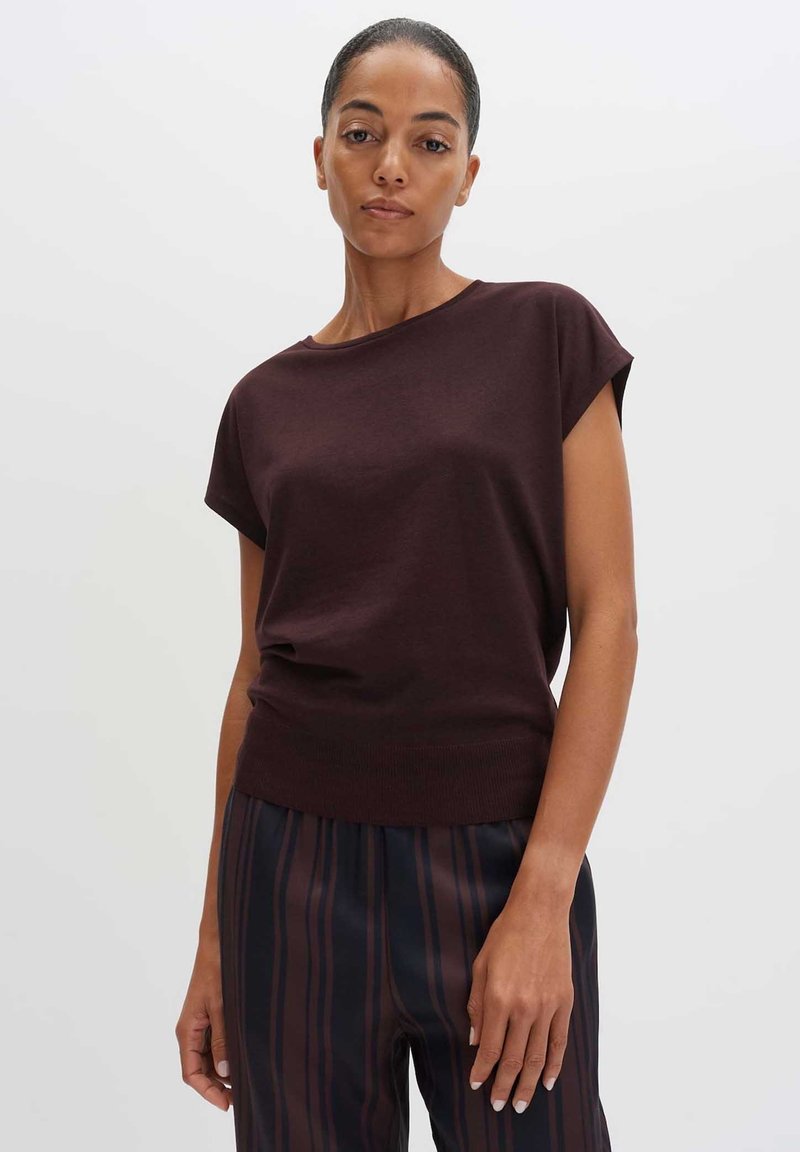 Woman with pulled-back hair wearing a dark brown short-sleeve top and black pants with vertical brown stripes, standing against a plain background.