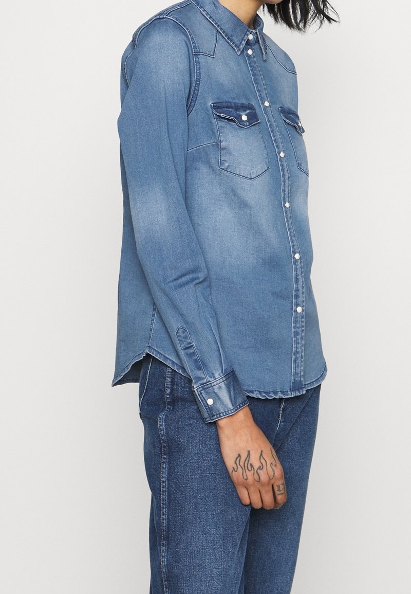 Ljusblå denimskjorta med spetsig krage, två bröstfickor och knappstängning. Långa ärmar med knäppta manschetter och rundad nederkant.