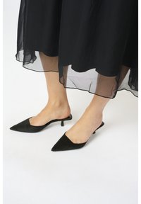 Mules noirs à talons hauts avec un bout pointu, dotés d'une tige en daim doux et d'un petit talon kitten, assortis à un ourlet de robe noire transparente.