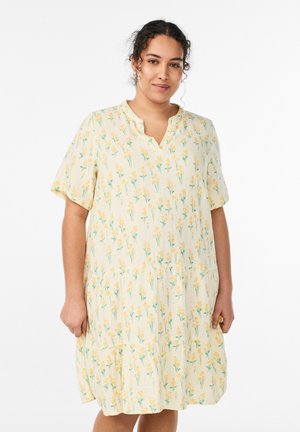 Kobieta o kręconych włosach, ubrana w luźną, kremową sukienkę z krótkim rękawem, ozdobioną małymi żółtymi kwiatami, stojąca na białym tle.