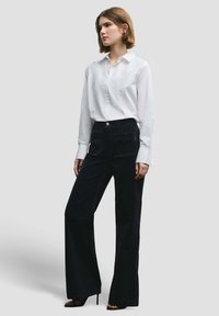 Wit overhemd met lange mouwen, gecombineerd met zwarte corduroy broek met hoge taille, voorzien van twee voorzakken en een wijde pijp.