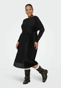 Robe noire à manches longues avec un corsage ajusté, une taille ceinturée et une jupe fluide. Portée avec des bottes noires jusqu'au genou et des chaussettes couleur crème.