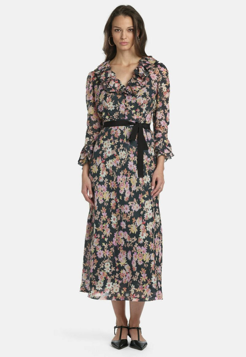 Femme portant une longue robe fleurie avec un col et des manches volantés, une ceinture noire à la taille et des chaussures noires à bout pointu, debout devant un fond blanc.