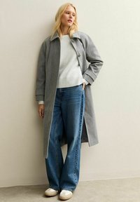 Femme blonde portant un long manteau gris par-dessus un pull léger, un jean bleu ample et des baskets blanches, adossée à un mur beige.