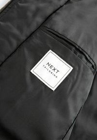 Musta räätälöity takki, jonka sisäpuolella on sileä tekstuuri, tyylikäs muotoilu, pieni tasku ja valkoinen lappu, jossa on "NEXT TAILORING" -logo.