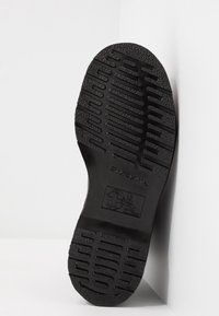 Semelle d'une chaussure noire avec motif de grip texturé et texte embossé, inclinée contre un mur d'angle blanc.