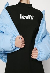 Giacca a vento blu chiaro con polsini elasticizzati, aperta sopra una maglietta nera con il logo bianco "levi's". Texture liscia e design moderno.