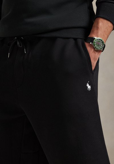 Μαύρο παντελόνι φόρμας με κορδόνι στη μέση, πλαϊνές τσέπες και μικρό λευκό λογότυπο. Συνοδεύεται από πράσινο ρολόι με υφασμένο λουράκι.