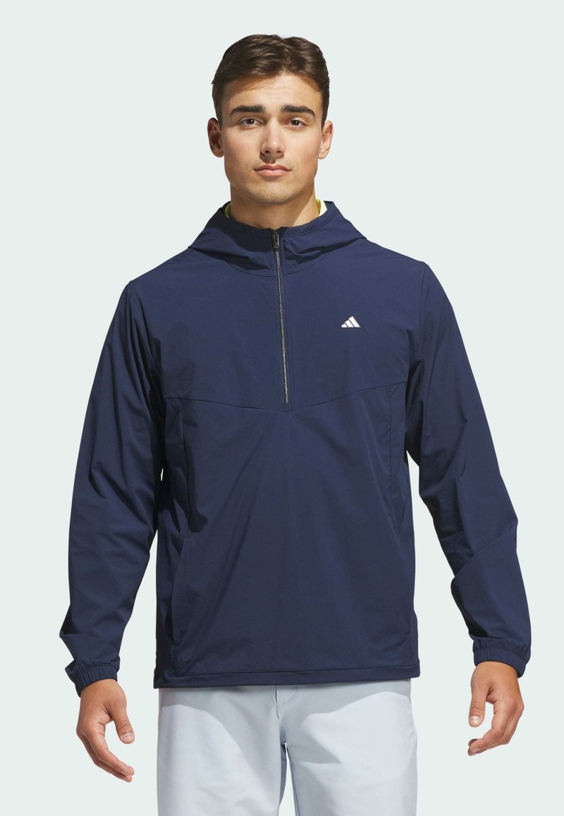 Homme portant un pull-over bleu marine à demi-fermeture éclair avec capuche et un pantalon blanc, debout devant un fond clair uni.