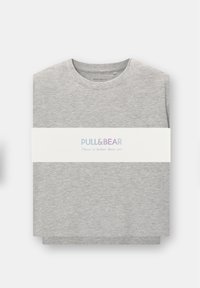 Szürke póló, normál fazonban, kerek nyakkal. Fehér szalaggal átcsomagolva, amelyen a "PULL&BEAR" felirat és színes szöveg, valamint egy szlogen található.