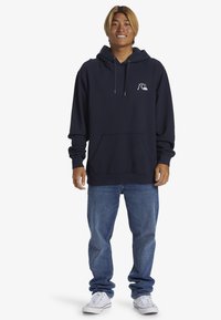 Felpa blu navy con tasca frontale, cappuccio con cordino e logo bianco. Abbinata a jeans azzurri e sneakers bianche. Look casual.