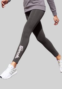 Donkergrijze leggings met een gladde textuur en een wit merklogo langs het linkerpijp. Gecombineerd met witte sport schoenen.