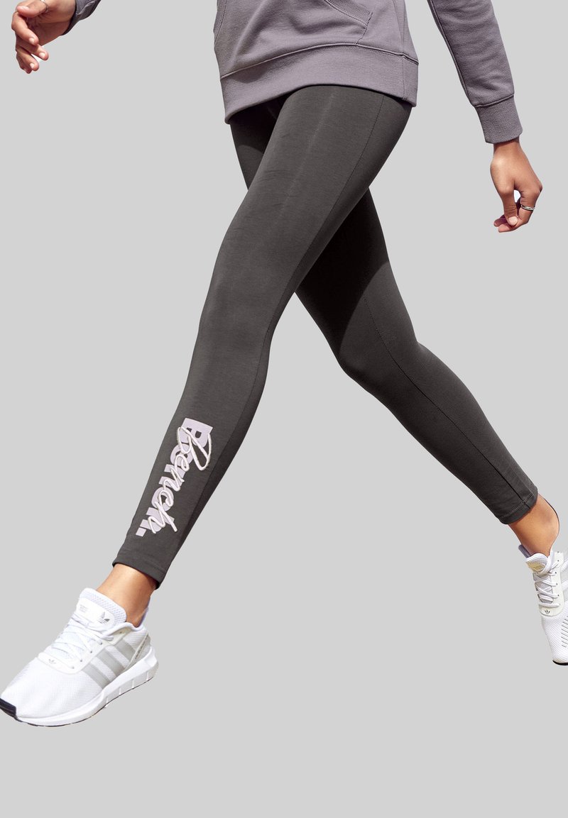 Donkergrijze leggings met een gladde textuur en een wit merklogo langs het linkerpijp. Gecombineerd met witte sport schoenen.