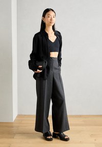Černá košile na knoflíky přes černý crop top, kombinována se šedými širokými kalhotami a černými platformovými sandály. Hladké materiály a moderní design.