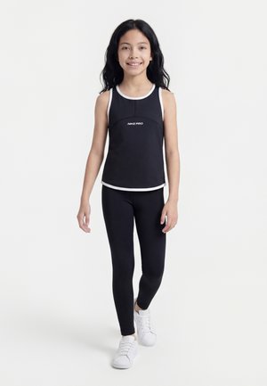 Mädchen geht, trägt ein schwarzes Nike Pro ärmelloses Top mit weißem Rand, schwarze Leggings und weiße Turnschuhe vor einem einfarbigen weißen Hintergrund.