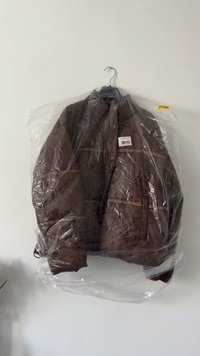 Bruine jas aan een zwarte hanger, bedekt met een doorzichtige plastic kledinghoes, hangend tegen een effen witte muur.