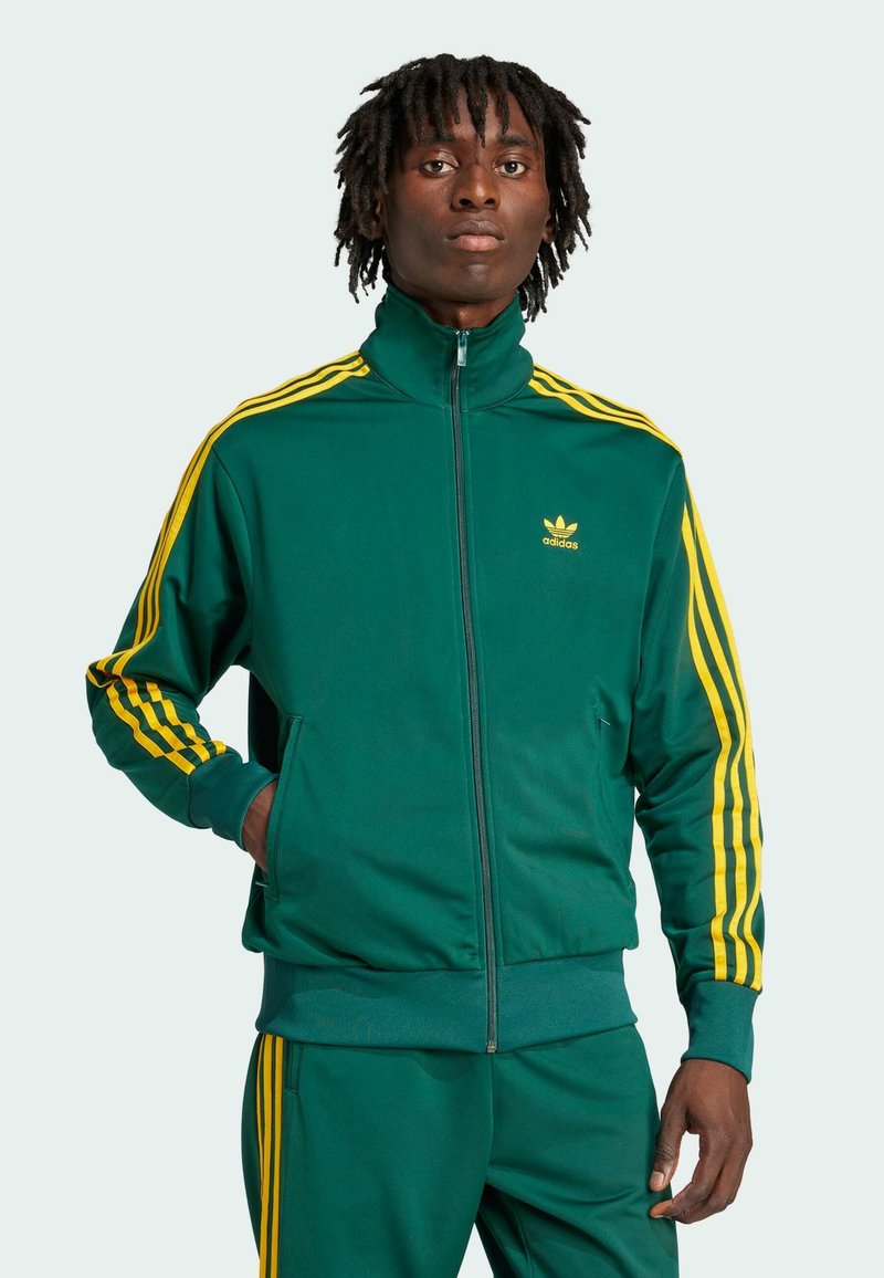 Zelená mikina se zipem a vysokým límcem vyrobená z hladkého materiálu, se žlutými třemi pruhy na rukávech a logem na hrudi.