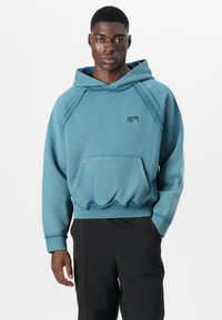 Blauer Baumwollkapuzenpullover mit einer Kängurutasche, verstellbarem Hoodie und Kontrastnähten. Mit einem Logo auf der Brust. Kombiniert mit schwarzen Hosen.