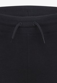 Un pantalon de survêtement noir présente une taille côtelée avec un cordon de serrage. Le tissu est lisse et a une couleur unie, sans motifs ni accents.