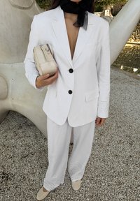 Valge linane bleiser mustade nööpidega, sama tooni laiade pükste komplekt, millel on silmadega aktsendid, ja väike beez kott. Must sall on seotud kaela ümber.