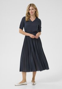 Mörkblå klänning med en plisserad, svävande kjol och en figurnära topp med V-ringning. Bärs med metalliska ballerinas.