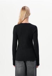 Blonde vrouw in een strakke zwarte lange mouwtop en losse vervaagde zwarte jeans, staand met haar rug naar de camera tegen een witte achtergrond.