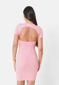 Rosa ribbet kjole med korte ærmer og åben ryg. Har en tætsiddende silhouette og brandtekst ved halsen. Glat tekstur.