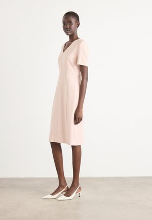 Robe rose blush, arrivant au genou, avec des manches courtes, un décolleté en V, à la coupe ajustée et des détails de couture, associée à des talons slingback blancs.