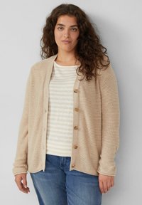 Beige gebreide cardigan met een textuurpatroon en houten knopen, gedragen over een gestreepte crèmekleurige en beige top, gecombineerd met blauwe jeans.