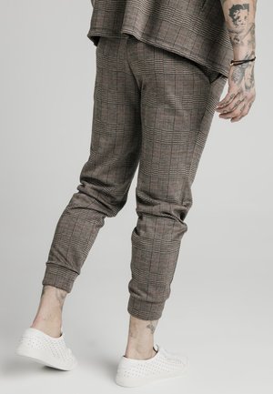 Homme portant un pantalon à carreaux marron et une veste assortie, des baskets blanches perforées et des tatouages visibles sur la main et la cheville.