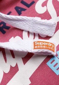 Tela rosa con letras blancas y azules. Es visible una etiqueta blanca que muestra la palabra "Superdry" en naranja con caracteres japoneses debajo.
