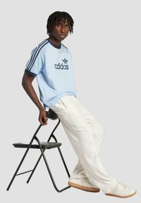Világoskék Adidas póló navy kiegészítőkkel, hálós textúrával és logóval; krémszínű textúrált nadrág; bézs sportcipő; fekete összecsukható szék.