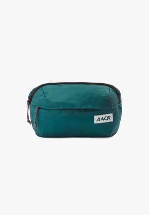 Pochette en tissu rectangulaire bleu sarcelle avec une fermeture éclair noire et des cordons, avec une petite étiquette de marque rectangulaire blanche à l'avant.