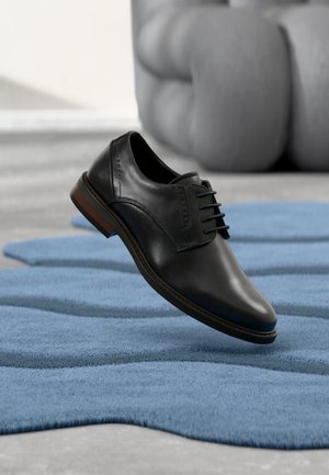 Zwarte leren veterschoen, geplaatst boven een blauw tapijt met golvend patroon, met op de achtergrond een grijze gestoffeerde bank.