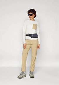 Fehér hosszú ujjú ing bézs zsebbel, bézs nadrág, és szürke túracipő. Fekete deréktáska és napszemüveg jellemzi.