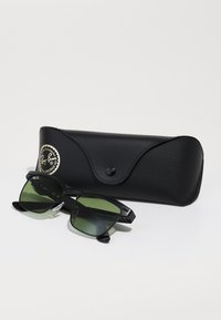 Ochelari de soare Ray-Ban negri cu lentile verzi, având o formă rectangulară și accente metalice, alături de o carcasă din piele neagră cu un logo.
