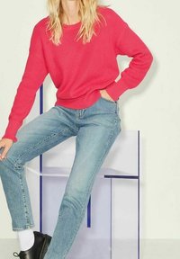 Maglione a coste rosa brillante abbinato a jeans azzurri, seduta su una sedia trasparente con un design geometrico.