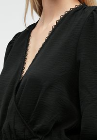 Musta mekko, jossa syvä V-pääntie, monimutkaiset pitsikoristeet, teksturoitu kangas ja puffy-hihat luovat virtaavan siluetin.