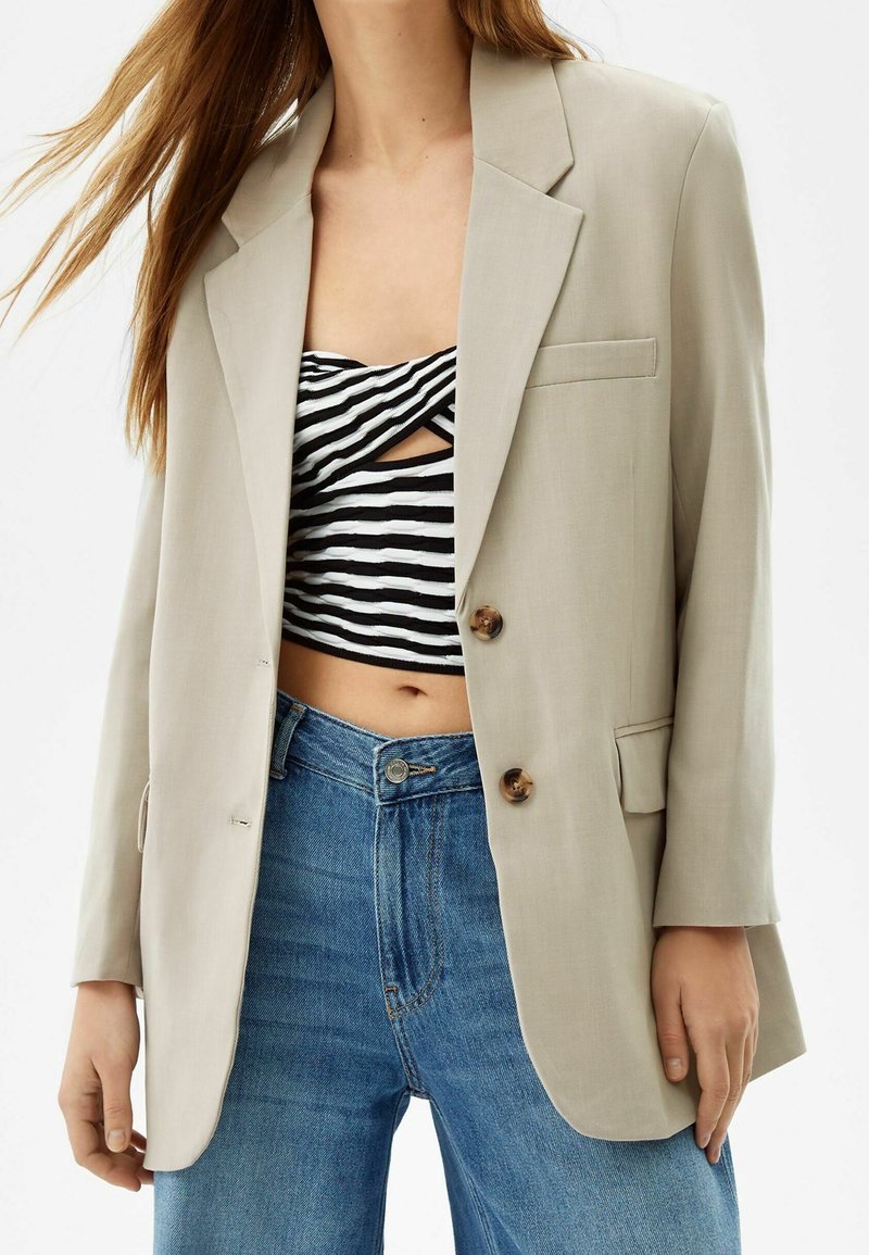 Femme portant un blazer beige oversize, un top court rayé noir et blanc, et un jean bleu taille haute sur un fond uni.
